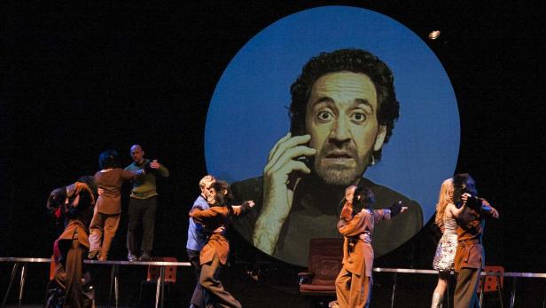 Eine Theaterszene mit Tänzern und einem großen projizierten Gesicht, das telefoniert.