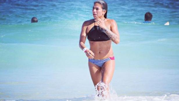 Eine Frau im Bikini watet durch das türkisfarbene Wasser am Strand.