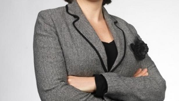 Eine Frau mit Brille und grauem Blazer posiert mit verschränkten Armen vor einem hellen Hintergrund.