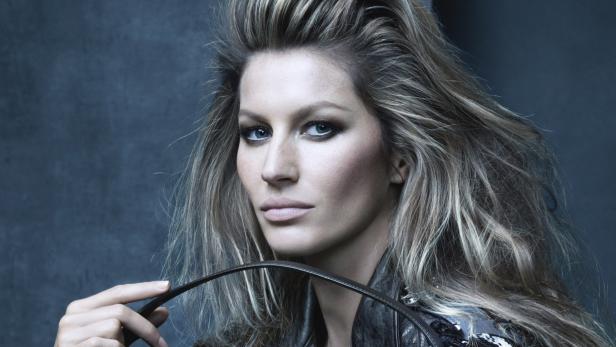 Gisele Bündchen mit starkem Make-up und Lederjacke vor dunklem Hintergrund.