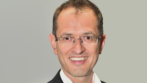 Ein lächelnder Mann mit Brille vor einem grauen Hintergrund.