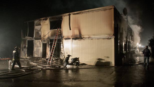 Ein zweistöckiges Gebäude steht in Flammen, während ein Feuerwehrmann und ein Polizist anwesend sind.