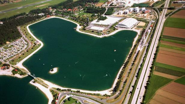 Luftaufnahme eines Sees mit umliegenden Gebäuden, Feldern und einer Autobahn.