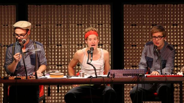 Drei Männer sitzen an einem Tisch mit Mikrofonen und Musikinstrumenten vor einem strukturierten Hintergrund.