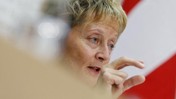 Eine Frau mit blonden Haaren gestikuliert vor einer rot-weißen Flagge.