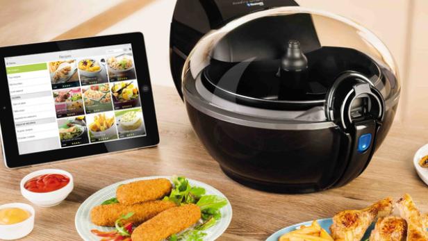 Eine Heißluftfritteuse der Marke Tefal steht neben einem Tablet und zwei Tellern mit frittierten Speisen.