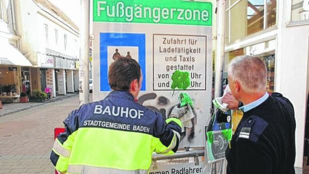 Zwei Bauhofmitarbeiter entfernen Graffiti von einem Schild in einer Fußgängerzone.