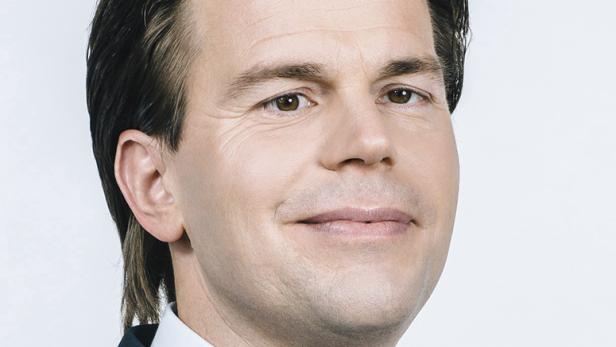 Porträt eines Mannes mit braunen Haaren und Anzug vor einem hellen Hintergrund.