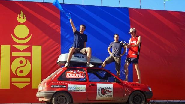 Drei Männer feiern auf einem roten Auto vor der Flagge der Mongolei die Mongol Rallye.