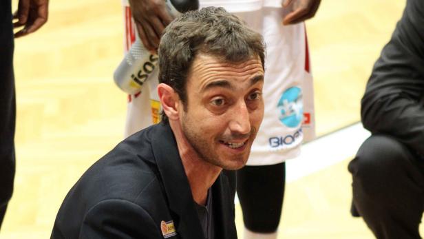 Ein Mann in einem schwarzen Anzug sitzt am Spielfeldrand eines Basketballspiels.