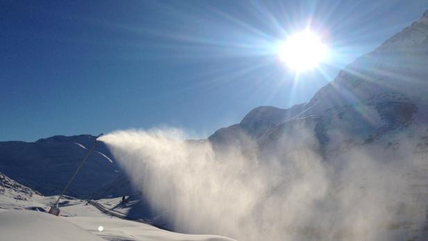 Eine Schneekanone erzeugt Kunstschnee vor einer Bergkulisse unter strahlendem Sonnenschein.