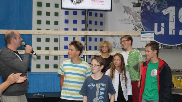 Eine Gruppe von Jugendlichen steht auf einer Bühne, während ein Mann ins Mikrofon spricht.