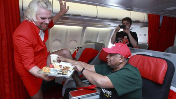 Richard Branson, verkleidet als Flugbegleiterin, serviert einem Passagier ein Tablett mit Speisen in einem Flugzeug.