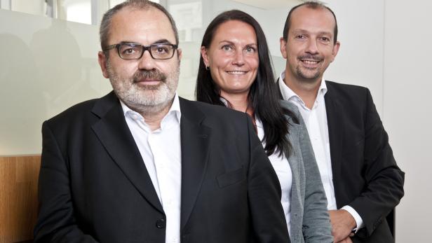 Drei Personen, zwei Männer in Anzügen und eine Frau, stehen nebeneinander und lächeln.