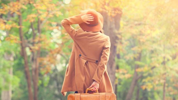 Eine Frau mit Hut und Koffer blickt in einem herbstlichen Park in die Ferne.