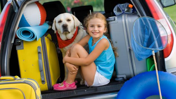 Ein Mädchen und ein Hund sitzen im Kofferraum eines Autos, umgeben von Reisegepäck und Strandutensilien.