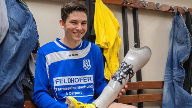 Ein lächelnder junger Mann im Fußballtrikot hält eine Beinprothese in der Hand.