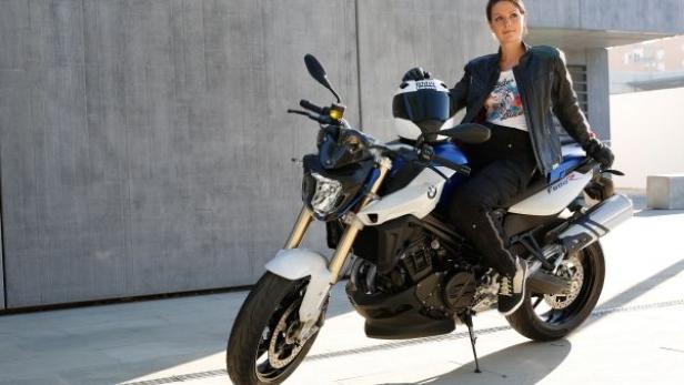 Eine Frau sitzt auf einem weißen BMW F800 R Motorrad vor einer grauen Wand.