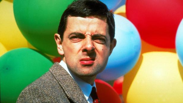 Rowan Atkinson als Mr. Bean mit zusammengekniffenem Gesicht vor bunten Ballons.