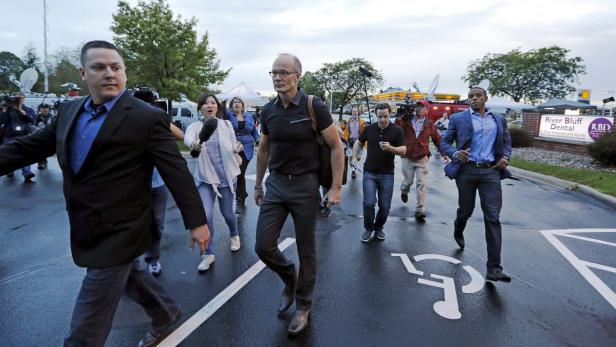 Eine Gruppe von Menschen, darunter Reporter mit Mikrofonen, geht auf einer nassen Straße.