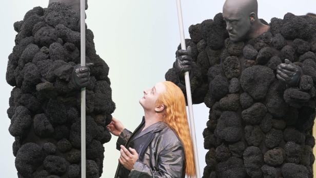 Eine Person mit roten Haaren interagiert mit zwei Wächtern in skulpturartigen, dunklen Kostümen.