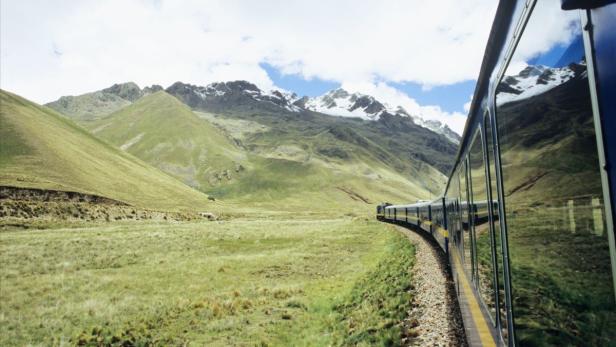 Ein Zug fährt durch eine grüne Landschaft mit Bergen im Hintergrund.