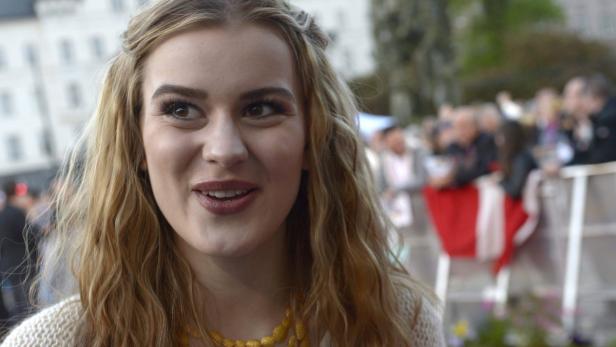 Eine junge Frau mit blonden, gewellten Haaren und einer goldenen Halskette lächelt.