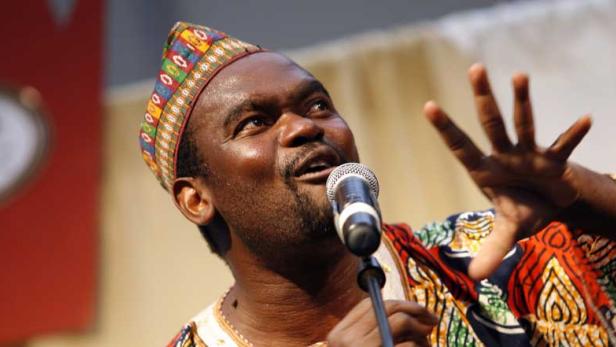 Ein Mann mit traditioneller Kopfbedeckung singt in ein Mikrofon und gestikuliert mit der Hand.