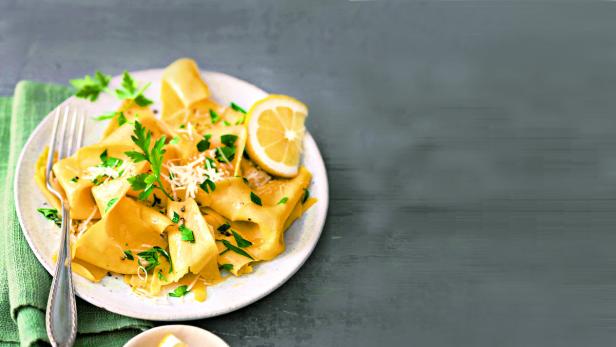 Ein Teller mit goldgelber Pasta, garniert mit Petersilie, Käse und einer Zitronenspalte.