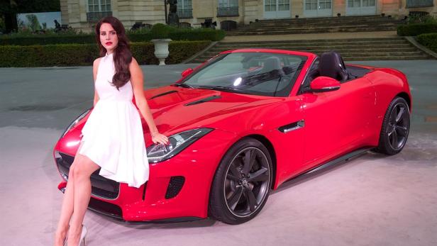 Lana Del Rey posiert an einem roten Jaguar F-Type Cabriolet.