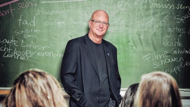 Ein Mann mit Brille steht vor einer Tafel, auf der Wörter wie „nette Lehrer“ und „Filme schauen“ stehen.
