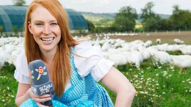 Eine rothaarige Frau mit Dirndl und Mikrofon steht vor einer Gänseherde.