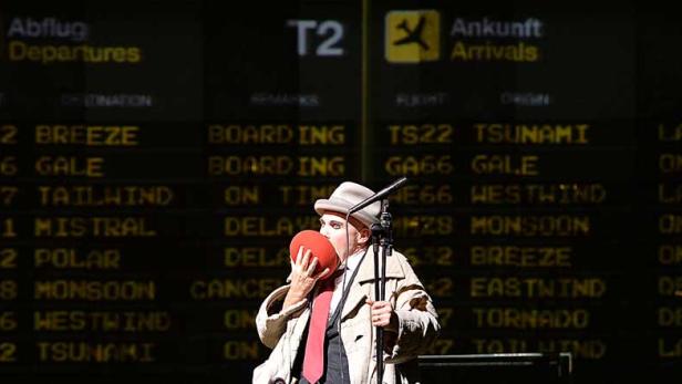 Ein Mann im Trenchcoat steht vor einer Anzeigetafel für Flugankünfte und -abflüge.