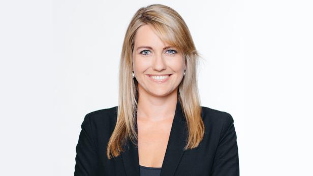 Eine blonde Frau mit blauen Augen lächelt in einem schwarzen Blazer.
