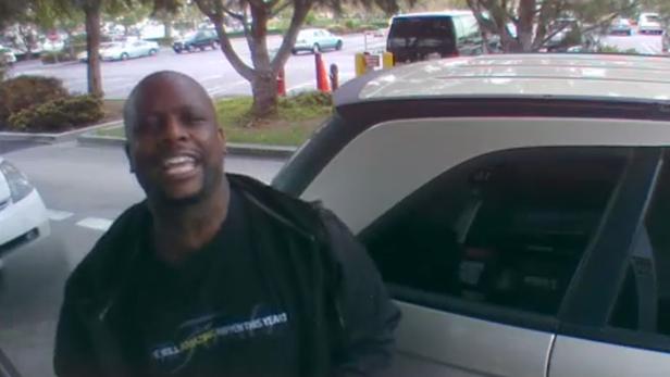 Ein lächelnder Mann steht vor einem Auto auf einem Parkplatz.