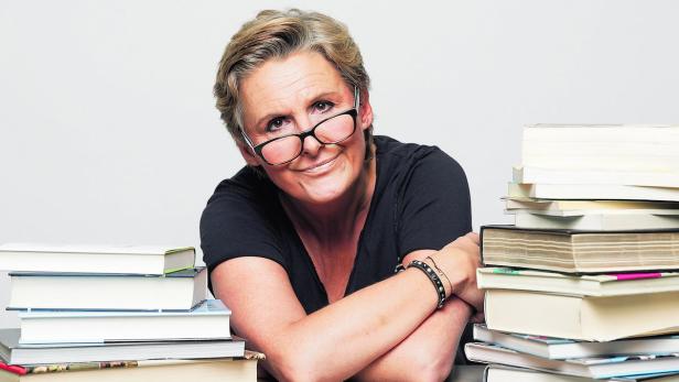 Eine Frau mit Brille sitzt lächelnd vor einem Stapel Bücher.