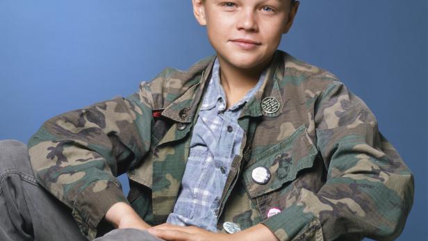 Ein junger Leonardo DiCaprio sitzt auf einem Stuhl vor einem blauen Hintergrund.