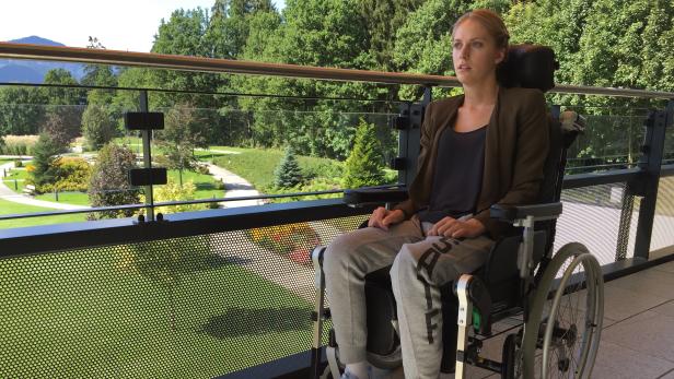 Eine junge Frau sitzt im Rollstuhl auf einem Balkon mit Blick auf einen Park.