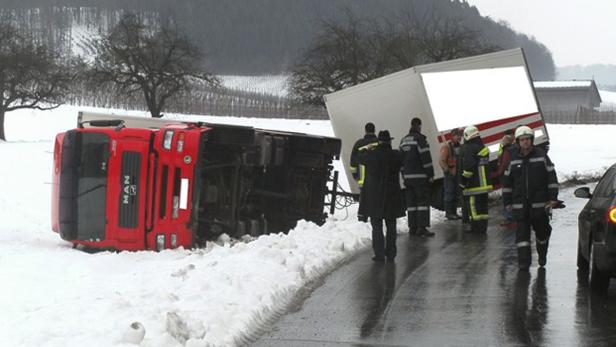 Ein umgekippter roter LKW liegt im Schnee, während Rettungskräfte am Unfallort stehen.