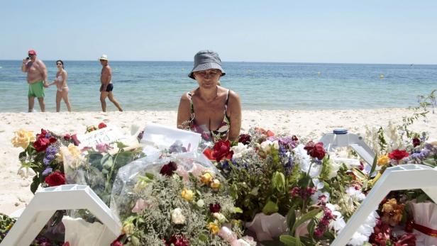 Eine Frau kniet am Strand vor einem Blumenmeer, im Hintergrund sind Badegäste zu sehen.