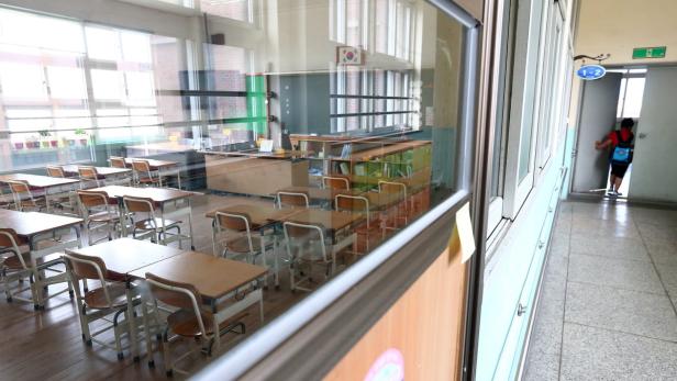 Blick durch ein Fenster in ein leeres Klassenzimmer mit Tischen und Stühlen. Eine Person geht den Flur entlang.