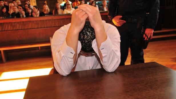 Ein Mann sitzt mit gesenktem Kopf an einem Tisch in einem Gerichtssaal, bewacht von einem Polizisten.