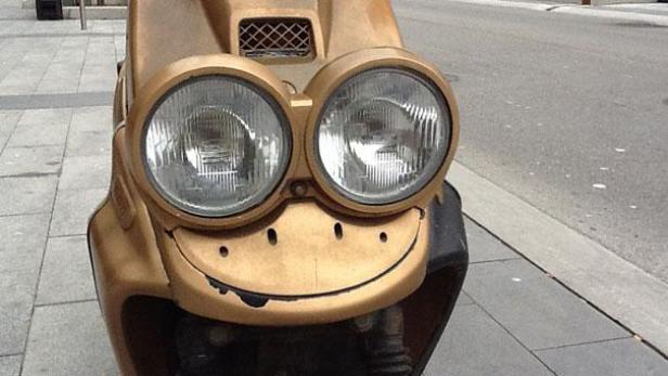 Ein goldener Motorroller mit aufgemaltem Gesicht steht am Straßenrand.