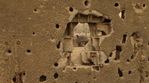 Ein Mann belädt einen Karren mit Metallschrott, gesehen durch ein Loch in einer zerstörten Mauer.