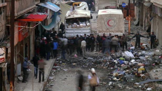 Menschen stehen vor Lastwagen des Roten Halbmonds in einer zerstörten Straße.