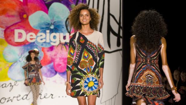 Eine lächelnde Frau präsentiert ein buntes Kleid von Desigual auf dem Laufsteg.