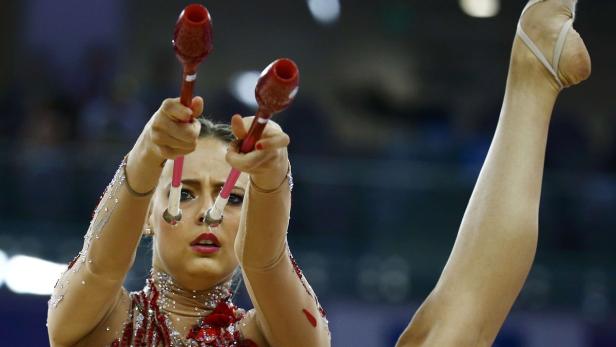 Eine rhythmische Sportgymnastin führt eine Übung mit Keulen vor, während sie ein Bein hochhebt.