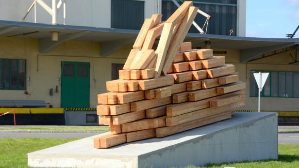 Eine Skulptur aus Holz auf einem Betonsockel vor einem gelben Gebäude.