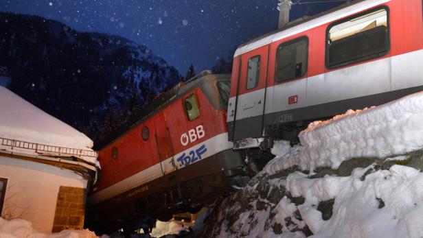 Zwei rote ÖBB-Züge sind in einen Unfall verwickelt und stehen teilweise auf einem schneebedeckten Hang.