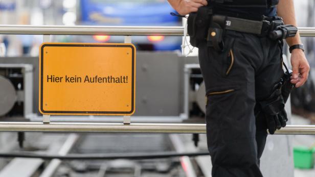 Ein gelbes Schild mit der Aufschrift „Hier kein Aufenthalt!“ steht neben einem Gleis und einem Polizisten.